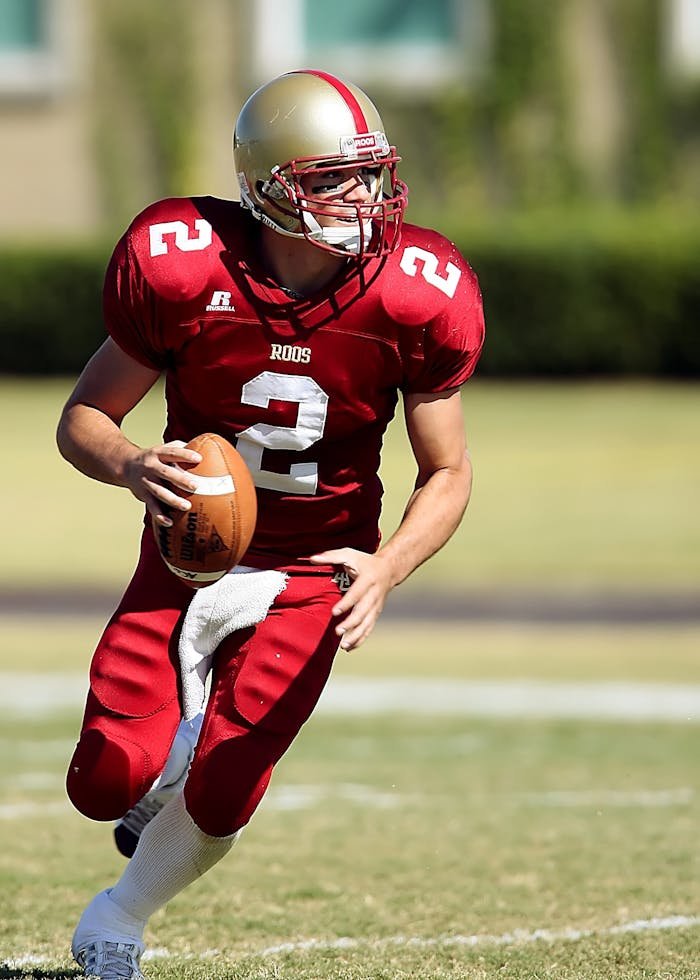 service-02 Quarterback in red jersey running with football on grassy field during daytime sporting event.