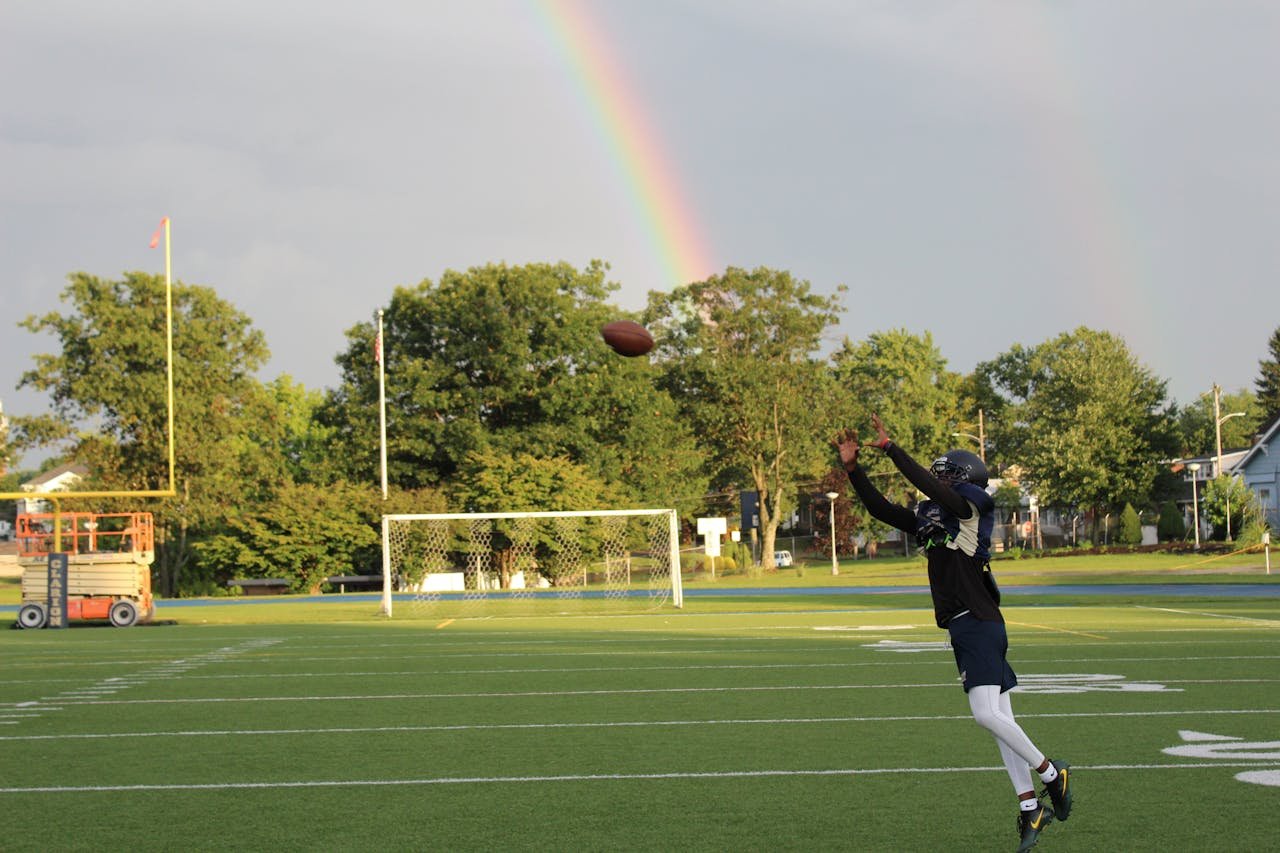 about-me Football player leaps to catch a ball with a vibrant rainbow backdrop on a sunny field.