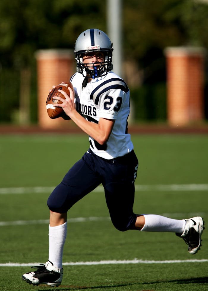 who-we-are Young quarterback running with a football during a game on a sunny day.